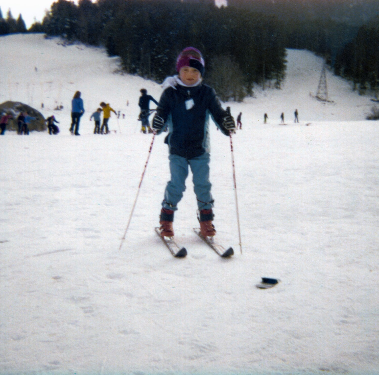 Photographie couleur de famille : un petit garçon à skis, en bonnet et anorak, les bâtons écartés sur une piste enneigée, d'autres skieurs et une forêt de sapins derrière lui — après restauration