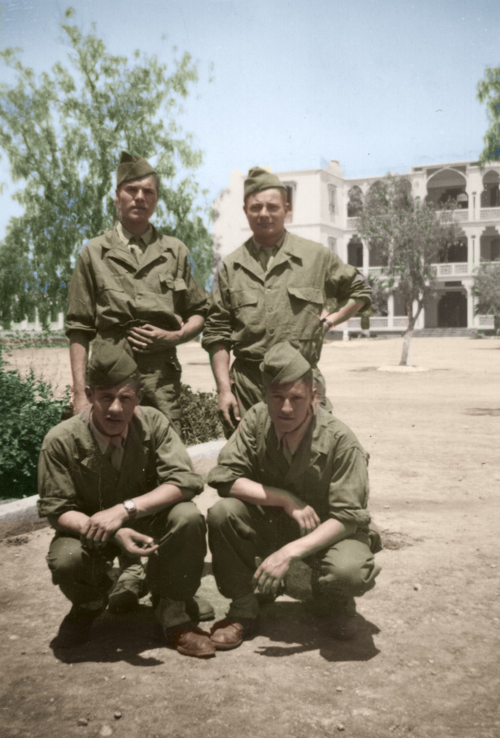 Quatre hommes en tenue militaire devant un grand bâtiment blanc à arcades, deux debout, deux accroupis — mai 1951 — restaurés et colorisés