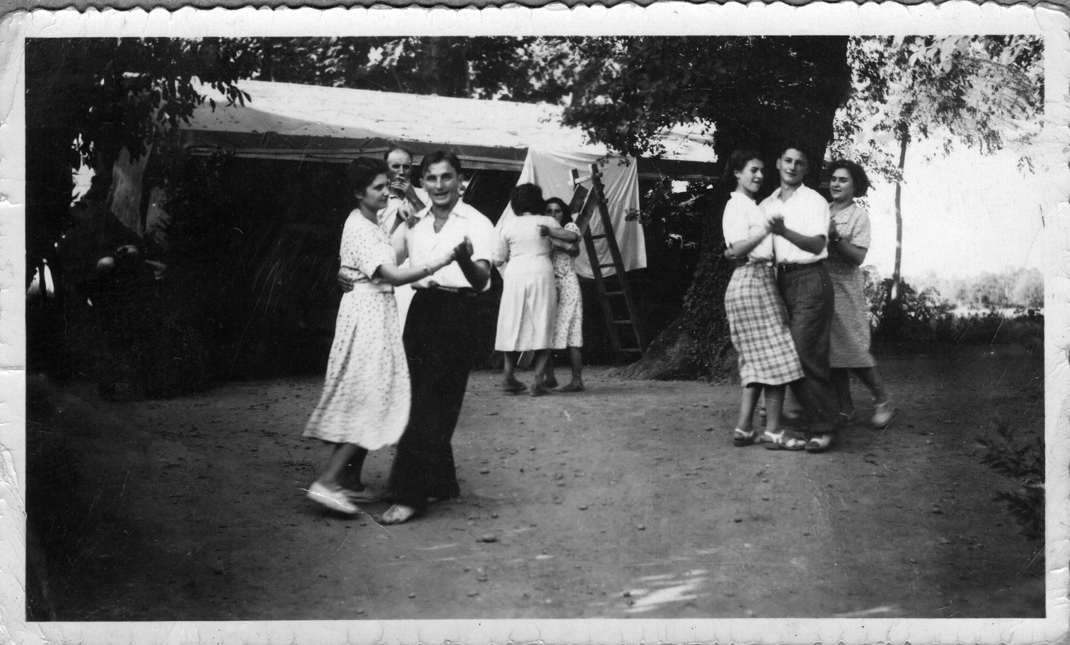 Photographie noir et blanc ancienne montrant plusieurs couples dansant dehors sous des arbres, avec du linge suspendu en arrière-plan et un bord festonné conservé — après restauration