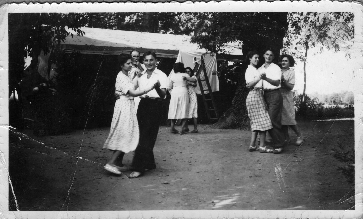 Photographie noir et blanc ancienne montrant plusieurs couples dansant dehors sous des arbres, avec du linge suspendu en arrière-plan et un bord festonné conservé — tirage d'origine, avant restauration
