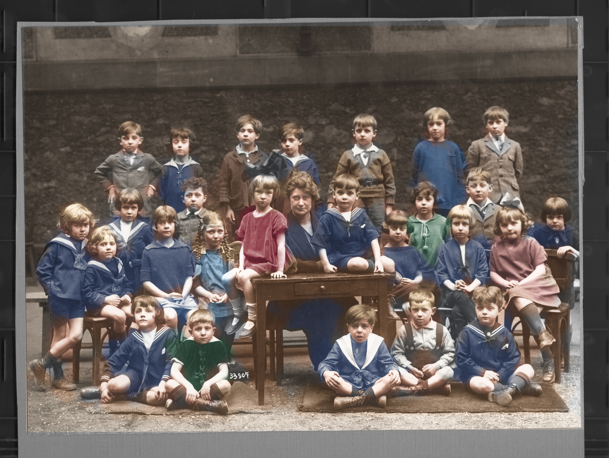 Version colorisée de la photographie de classe « Rigueur scolaire », 1922–1923 : vingt-six enfants et leur maîtresse posent dans une cour, autour d'une table d'école. Les couleurs sont une interprétation.