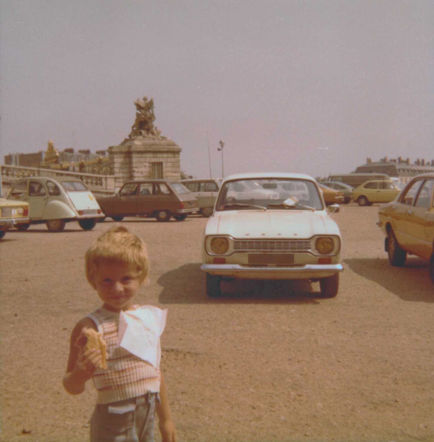 Photographie couleur de famille : un petit garçon, un gâteau à la main, devant une Ford blanche et d'autres voitures garées ; derrière, une statue sur son pilier et la grille dorée du château de Versailles, sous un ciel gris voilé — après restauration