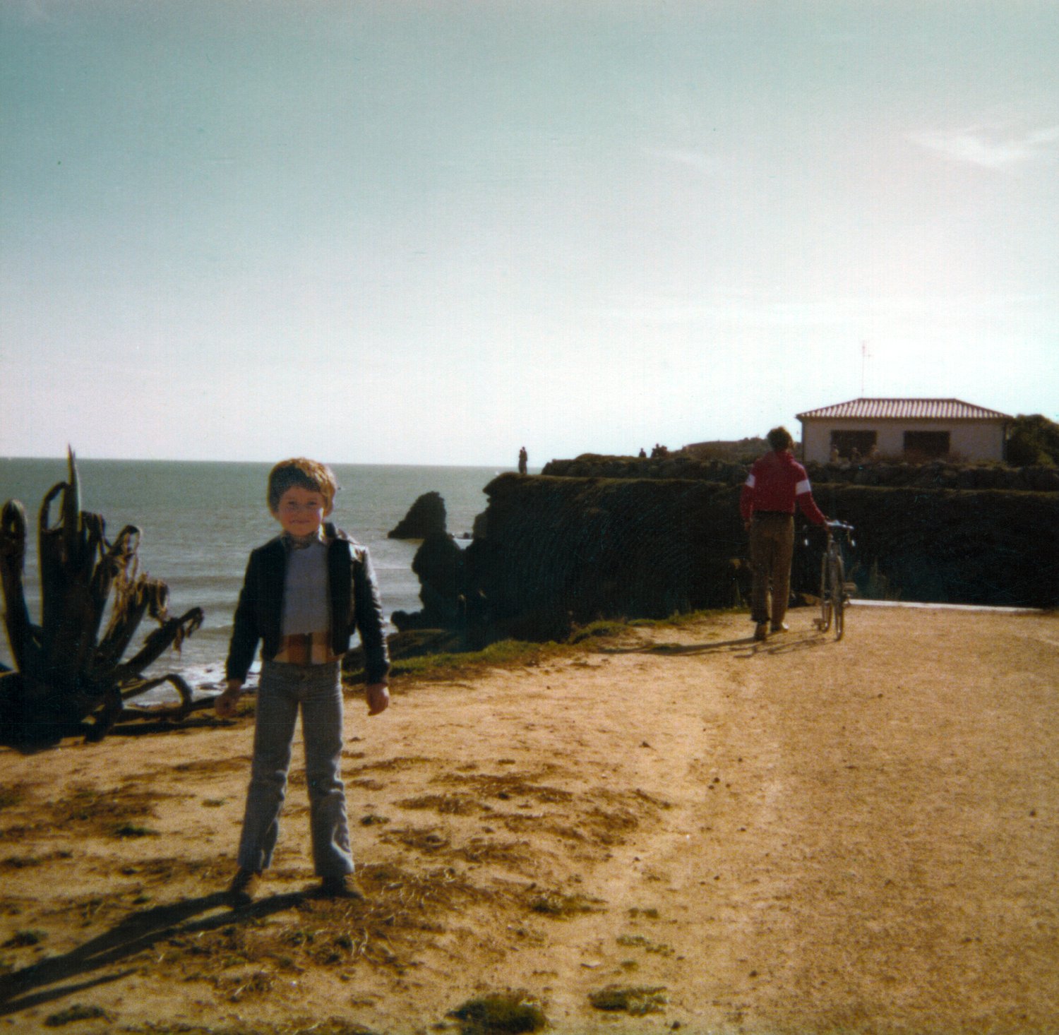 Photographie couleur de famille : un petit garçon en blouson sur un chemin au bord de la mer, une falaise et une maison blanche au loin, ciel pâle — après restauration